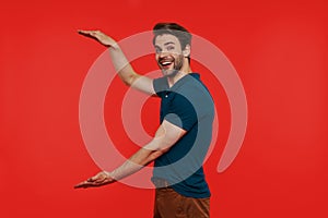 Happy young man in casual wear looking at camera and gesturing while standing against red background