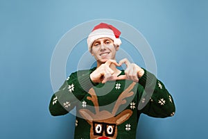 Happy young man in christmas sweater and hat santa stands on a blue background and shows in his hands a gesture of Heart. Cheerful