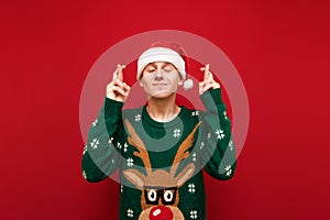 Happy young man in christmas sweater and santa hat makes a wish for christmas with closed eyes on red background, smiling.
