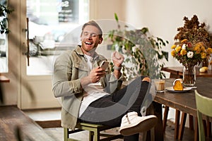 Happy young man in coffee shop, drinking cappuccino, listening to funny podcast in earphones, holding smartphone