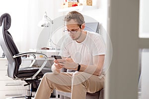 Happy young man in eyeglasses sitting on sofa sending text on mobile phone