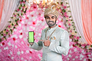 Happy young Man or Groom in Ethnic Weddding wear or attire showing green screen mobile phone on mariage stage - concept