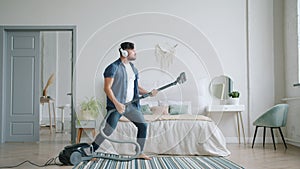 Happy young man in headphones singing in vacuum cleaner dancing cleaning home