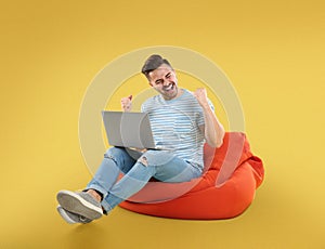 Happy young man with laptop on bean bag chair, yellow background