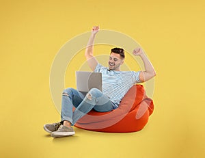 Happy young man with laptop on bean bag chair, yellow background