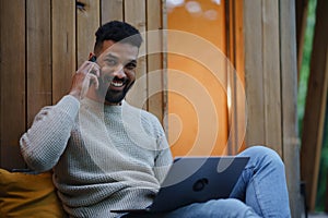 Happy young man with laptop and smartphone resting outdoors in a tree house, weekend away and remote office concept