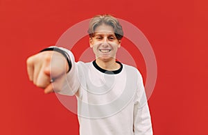 Happy young man in light casual clothes shows his finger to the camera with a smile on his face on a red wall background