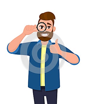 Happy young man looking through magnifier and showing thumbs up gesture sign. Trendy person holding magnifying glass or lens.