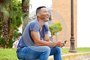 Happy young man with mobile phone sitting on sidewalk and looking away