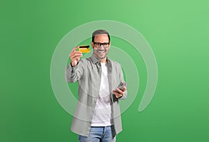 Happy young man with phone showing credit card and recommending online payment on green background