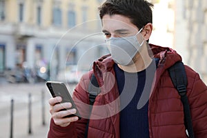 Happy young man with protective mask looks his smartphone in street. Student guy with face mask using mobile phone outdoors