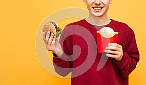 Happy young man in red sweatshirt with burger and fries in hands, smiling, isolated on yellow background, cropped close up photo.