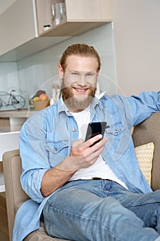 Happy young man relaxing on sofa using smartphone looking at camera at home.