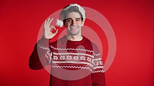 Happy young man in Santa hat showing Ok sign gesture isolated on red