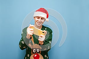 Happy young man in santa hat and warm cut-up sweater plays mobile games against a blue background, looks into the camera and