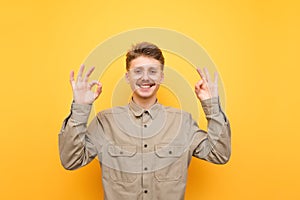 Happy young man in shirt and glasses smiling, looking at camera and showing thumbs up gesture OK on yellow background. Portrait of