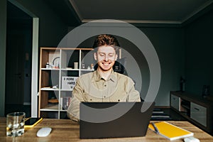 Happy young man in shirt working at home on laptop with smile on face. Joyful freelancer works at home on remote work. The guy