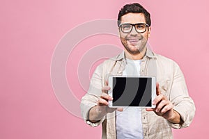 Happy young man showing blank tablet computer screen and looking at the camera isolated over pink background