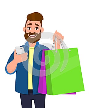 Happy young man showing shopping bags. Person holding a mobile, cell, smart phone in hand. Modern lifestyle, digital technology.
