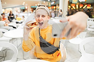 Happy young man sitting at fast food restaurant eating burger and taking selfie. A guy in casual clothes poses on a smartphone