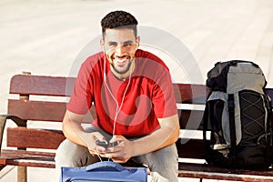 Happy young man sitting with luggage and listening to music with mobile phone