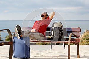 Happy young man sitting outside with luggage and listening to music with mobile phone and earphones