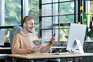 A happy young man is sitting at the table in the lobby, working at the computer. Holds cash, dollars and uses the phone