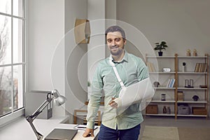 Happy young man with a sling on his broken arm standing by his working desk at home