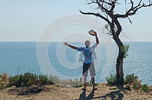 Happy young man standing on a cliff with his arms raised looks at the sea