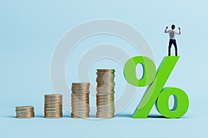 Happy young man standing on growing stacked golden coins and green interest rates on blue background. Financial business concept,