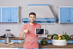 Happy young man standing in kitchen and pointing at tablet, looking at camera with toothy smile