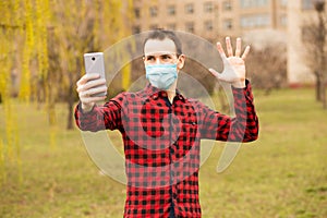 Happy young man in sterile face mask making video call with mobile phone show hello gesture walking by street