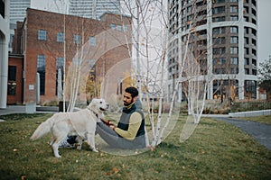 Happy young man stroking his dog outdoors in city park, during cold autumn day.