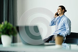 Happy young man talking on cell phone and using laptop in office