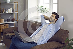 Happy young man thinking and dreaming while relaxing on comfortable couch with laptop