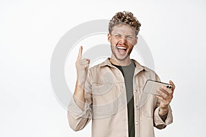 Happy young man triumphing, holding mobile phone and showing rock n roll, heavy metal gesture and shouting joyful. Guy
