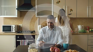 Happy young man using digital tablet computer sitting in the kitchen while his girlfriend cooking at home