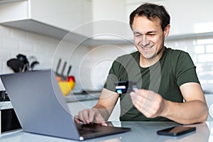 Happy young man using laptop computer while sitting at the kitchen table use credit card for online purchase
