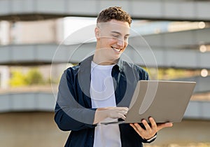 A happy young man using a laptop outdoors, dressed in a casual