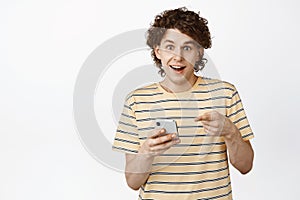 Happy young man using mobile phone and smiling, pointing at his screen, receive an announcement on smartphone, standing