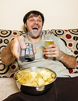 Happy young man watching television, eating potato chips and drinking beer