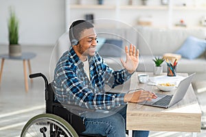Happy young man in wheelchair communicating online, using laptop computer, waving at webcam at home