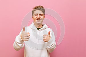Happy young man in white casual clothes, looks into the camera with a smile on his face and shows thumbs up, isolated on pink