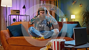 Happy young man in wireless headphones relaxing sitting on sofa at home listening favorite music