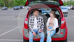 Happy young man and woman sitting on trunk of car outdoors while talking. Transportation, leisure, road trip, travel and