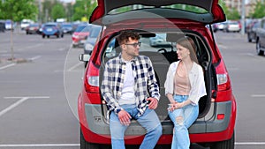 Happy young man and woman sitting on trunk of car outdoors while talking. Transportation, leisure, road trip, travel and