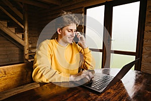 Happy young man working on laptop in the country house in a room with wooden interior and talking on the phone with a smile on his