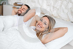 Happy young married couple relaxing on soft pillows in bed, enjoying lazy weekend morning at home, copy space