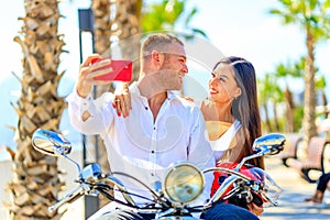 Happy young married couple on a scooter in honeymoon travel in paradise beach taking selfie photo on smartphone camera