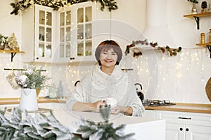 Happy young middle aged woman enjoying drinking tea or coffee in kitchen with Christmas decor